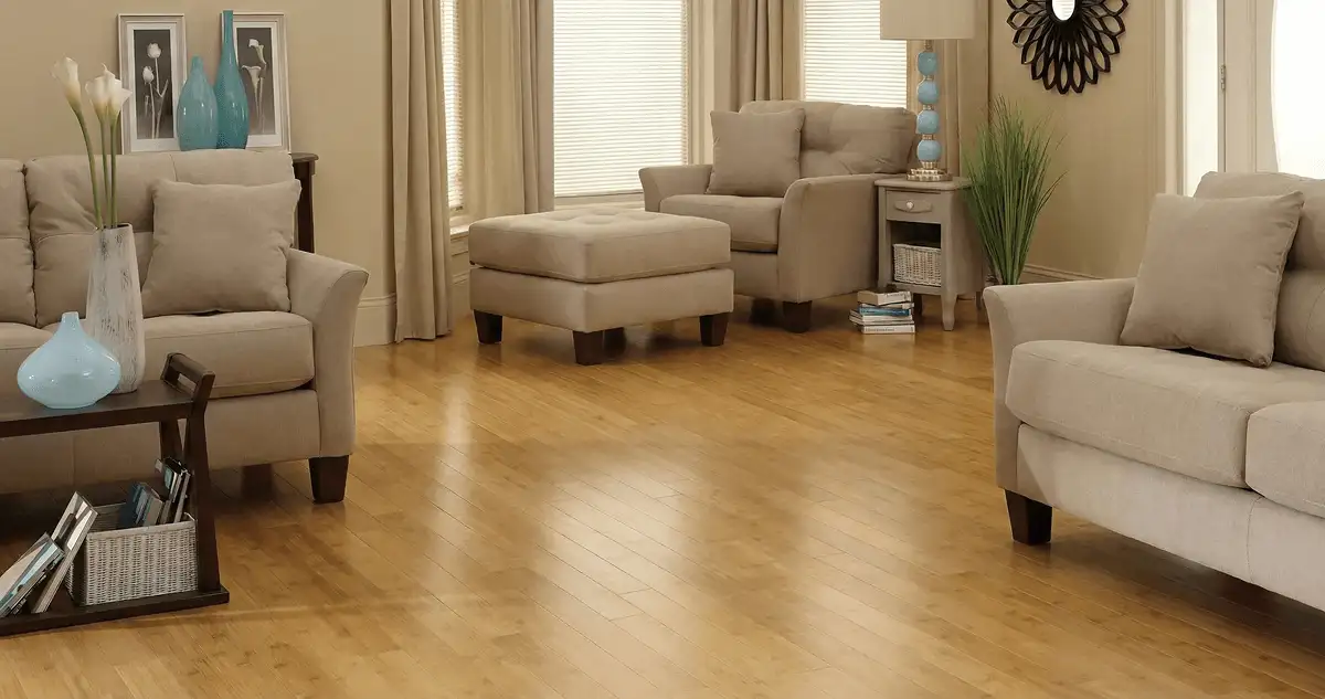A living room with professionally installed bamboo flooring, beige sofas, a matching ottoman, a side table with a blue lamp, vases, framed pictures, and large windows with blinds and curtains in Colorado Springs, CO